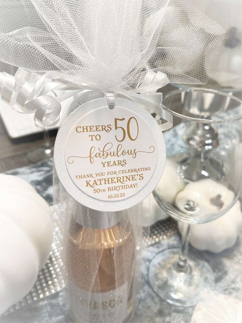 May include: White round favor tag with gold lettering. It says "Cheers to 50 fabulous years. Thank you for celebrating Katherine's 50th Birthday! 10.22.22".