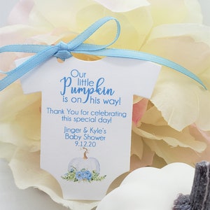May include: A white baby onesie shaped tag with blue ribbon. The tag says "Our little pumpkin is on his way! Thank You for celebrating this special day! Jinger & Kyle's Baby Shower 9.12.20". The tag has a watercolor illustration of a white pumpkin with blue flowers.