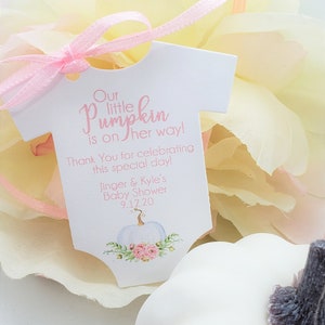 May include: A white baby onesie shaped tag with pink ribbon. The tag says "Our little pumpkin is on her way! Thank You for celebrating this special day! Jinger & Kyle's Baby Shower 9.12.20". The tag has a white pumpkin with pink flowers and green leaves.