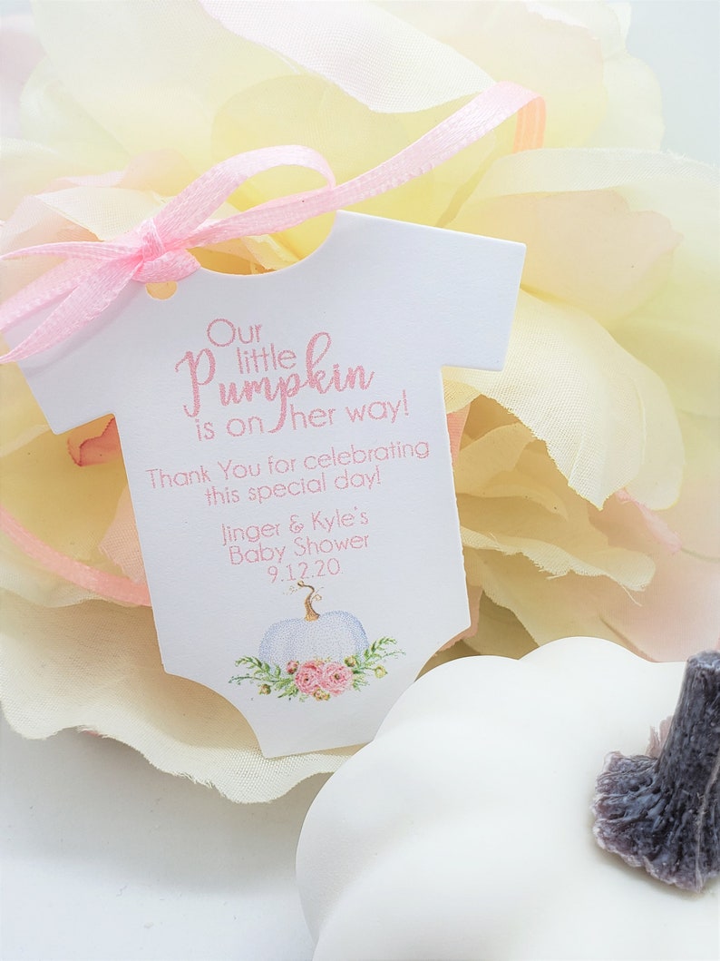 May include: A white baby onesie shaped tag with pink ribbon. The tag says "Our little pumpkin is on her way! Thank You for celebrating this special day! Jinger & Kyle's Baby Shower 9.12.20". The tag has a white pumpkin with pink flowers and green leaves.