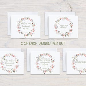 May include: Set of white greeting cards with floral wreath designs. Each card features a different inspirational quote in elegant script. The wreaths are adorned with pink and white flowers and green leaves. The text "2 OF EACH DESIGN PER SET" is also visible.