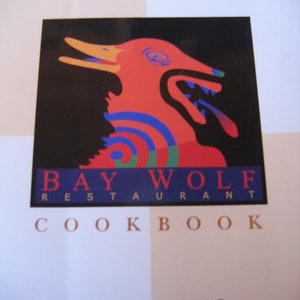 May include: A cookbook cover featuring a stylized red wolf illustration with blue, green, and orange accents against a black background. The text reads "BAY WOLF RESTAURANT COOKBOOK" with author names below.