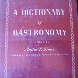 May include: A Dictionary of Gastronomy book cover. The cover is a deep red with white text and a decorative white border. The title reads "A Dictionary of Gastronomy" with the author's name, Andre L. Simon, and his title below.