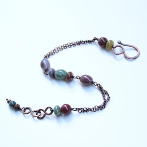 May include: A bracelet featuring a dark copper chain with a hook clasp. The design incorporates smooth, polished, multi-colored stone beads in shades of green, brown, and grey, interspersed along the chain links in an asymmetrical arrangement.
