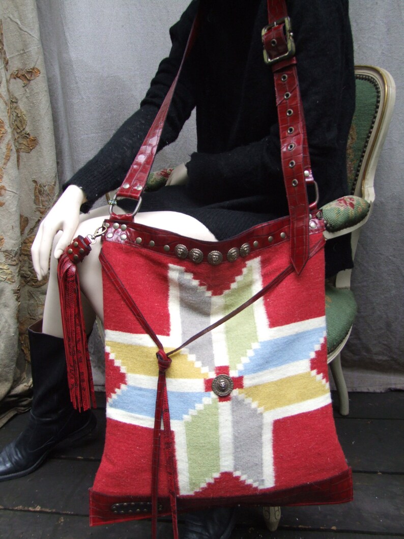 May include: A red, white, and blue woven tapestry bag with leather straps and metal accents. The bag has a geometric pattern and is worn over the shoulder.