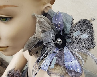 May include: A gray, black, and purple fabric collar with a large rhinestone embellishment. The collar is made of various textures and fabrics, including lace, tulle, and felt.