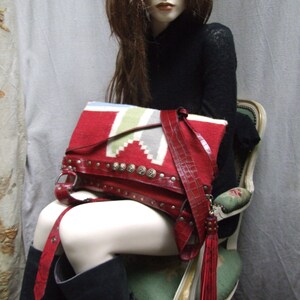 May include: A red leather bag with a fringe tassel. The bag has a woven pattern and is decorated with metal studs. The bag is being held by a mannequin wearing a black sweater.