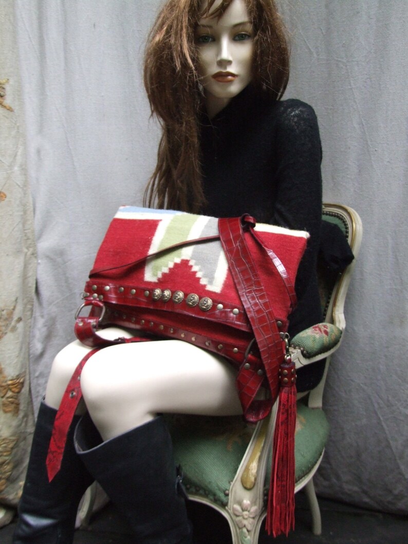May include: A red leather bag with a fringe tassel. The bag has a woven pattern and is decorated with metal studs. The bag is being held by a mannequin wearing a black sweater.