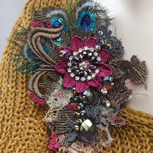 May include: A large, ornate brooch with a pink flower, peacock feathers, and iridescent beads. The brooch is attached to a brown knitted sweater.