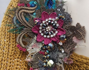 May include: A large, ornate brooch with a pink flower, peacock feathers, and iridescent beads. The brooch is attached to a brown knitted sweater.