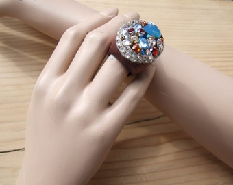 May include: A large, round ring with a brown base and a colorful assortment of rhinestones and glass stones. The stones are in shades of blue, red, orange, and clear.
