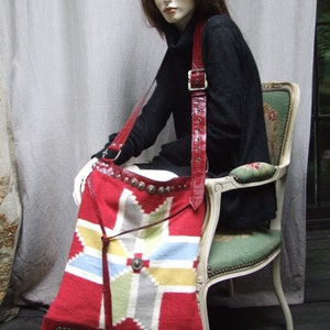 May include: A red and white woven tapestry bag with a leather strap and silver accents. The bag is worn by a mannequin wearing a black turtleneck sweater.