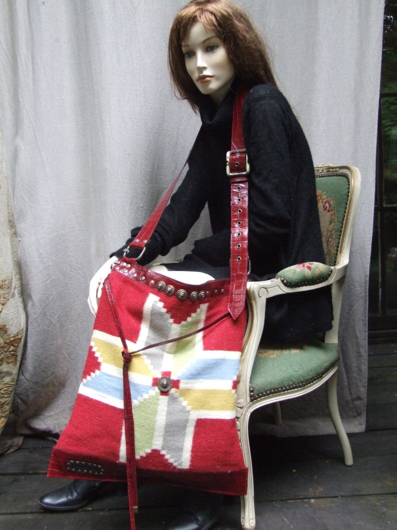 May include: A red and white woven tapestry bag with a leather strap and silver accents. The bag is worn by a mannequin wearing a black turtleneck sweater.