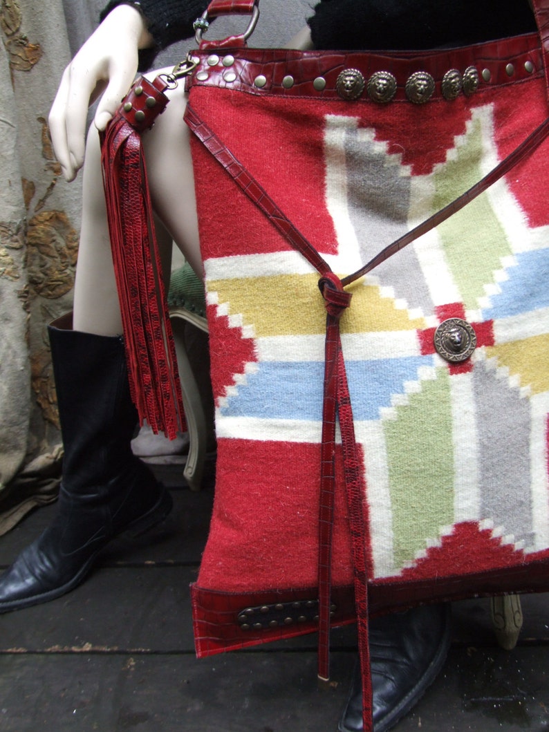 May include: A red, white, and blue patterned leather bag with a long fringe. The bag has a leather strap and metal accents.