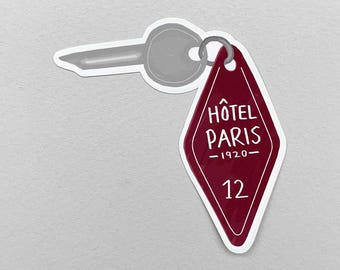Può includere: Un portachiavi da hotel bordeaux con le parole "HÔTEL PARIS -1920-" e il numero "12" in bianco. L'etichetta è a forma di diamante e attaccata a una chiave grigia. Lo sfondo è grigio chiaro.