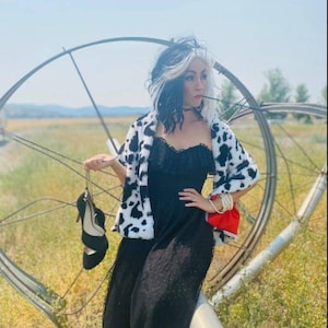 May include: A woman dressed as Cruella de Vil from the Disney movie 101 Dalmatians. She is wearing a black dress with a white and black dalmatian print fur coat. She is holding a black high heel shoe in her hand.