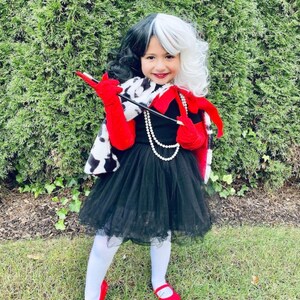 Pu&ograve; includere: Una bambina vestita da Cruella de Vil per Halloween. Indossa un cappotto con stampa dalmata bianco e nero, un vestito rosso e nero, guanti rossi e una parrucca bianca e nera. Tiene in mano un porta sigarette nero.