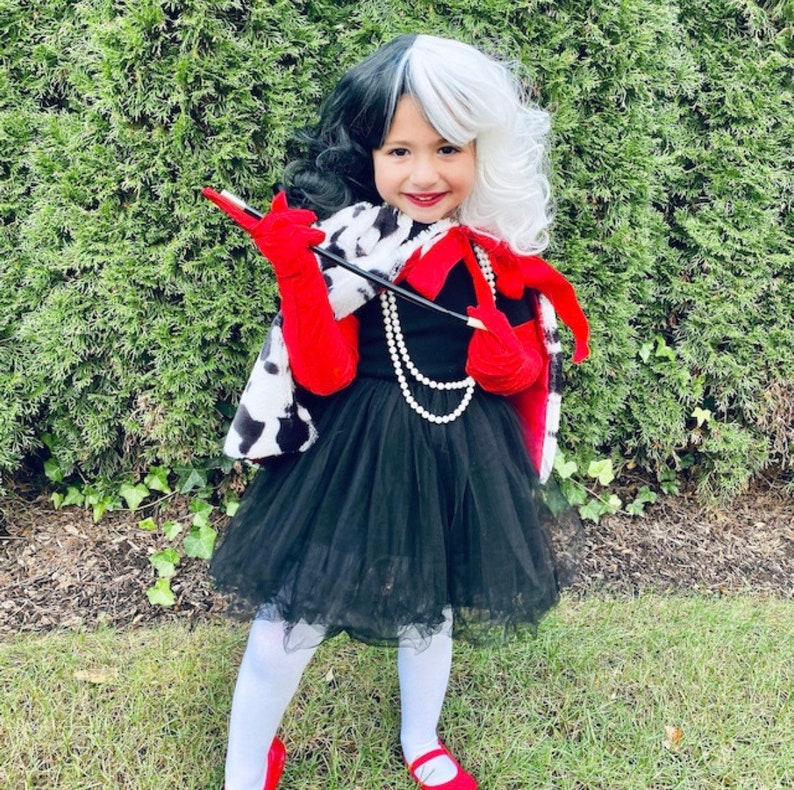 Pu&ograve; includere: Una bambina vestita da Cruella de Vil per Halloween. Indossa un cappotto con stampa dalmata bianco e nero, un vestito rosso e nero, guanti rossi e una parrucca bianca e nera. Tiene in mano un porta sigarette nero.