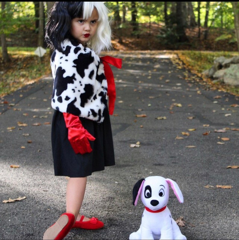 Pu&ograve; includere: Una bambina vestita come Cruella de Vil per Halloween. Indossa un cappotto con stampa dalmata bianco e nero, un vestito nero, guanti rossi e scarpe rosse. Ha una parrucca bianca e nera e rossetto rosso. &Egrave; in piedi accanto a un peluche dalmata bianco.