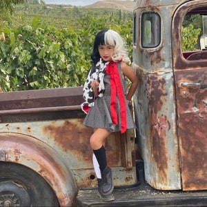 Pu&ograve; includere: Una giovane persona vestita come Cruella de Vil, con un cappotto a stampa dalmata bianco e nero, un vestito a pois rosso e nero e calze alte bianche e nere. &Egrave; in piedi accanto a un camion vintage arrugginito.