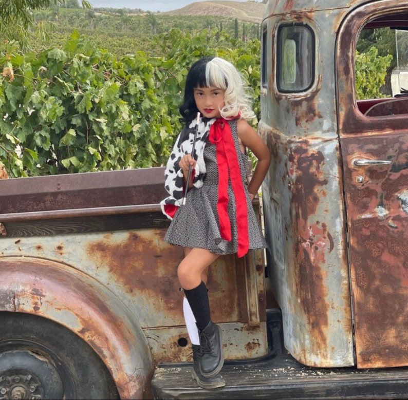 Pu&ograve; includere: Una giovane persona vestita come Cruella de Vil, con un cappotto a stampa dalmata bianco e nero, un vestito a pois rosso e nero e calze alte bianche e nere. &Egrave; in piedi accanto a un camion vintage arrugginito.