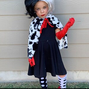 Pu&ograve; includere: Una bambina vestita da Cruella de Vil per Halloween. Indossa un vestito nero con un mantello a stampa dalmata, guanti rossi e calze a pois bianche e nere.