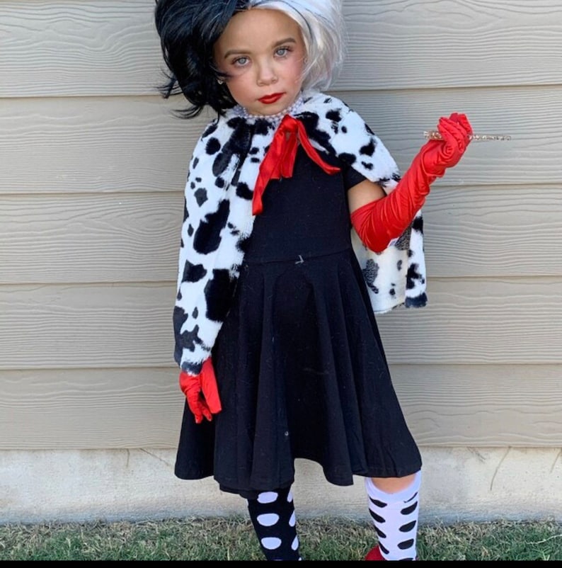 Pu&ograve; includere: Una bambina vestita da Cruella de Vil per Halloween. Indossa un vestito nero con un mantello a stampa dalmata, guanti rossi e calze a pois bianche e nere.