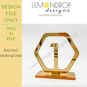 Puede incluir: Número de mesa uno en acrílico dorado de espejo en forma hexagonal. El número está en negrita y está sobre una base dorada.