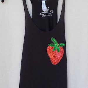 May include: Black tank top with a sequined strawberry design. The tank top has a racerback style and the text "Whi Creations" is printed on the back.