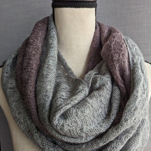 K&ouml;nnte beinhalten: Ein grauer und lilafarbener Infinity-Schal aus weichem, gestricktem Stoff. Der Schal ist um den Hals einer Schaufensterpuppe gelegt.