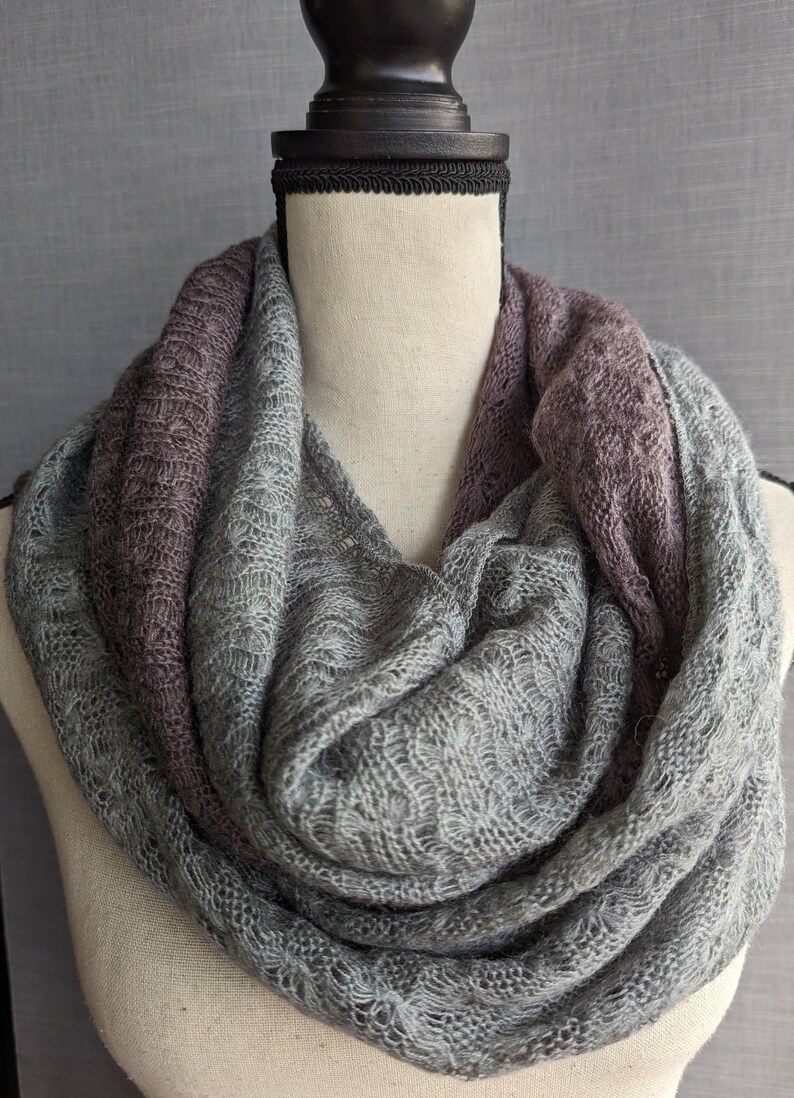 K&ouml;nnte beinhalten: Ein grauer und lilafarbener Infinity-Schal aus weichem, gestricktem Stoff. Der Schal ist um den Hals einer Schaufensterpuppe gelegt.