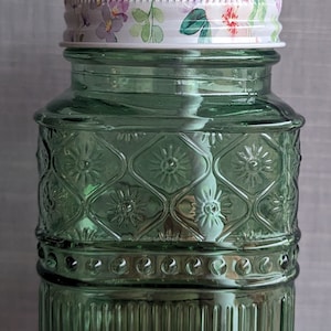 May include: A green glass jar with a floral patterned lid and a white straw. The jar has a textured design with a raised pattern of flowers and circles.