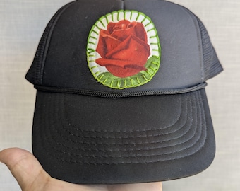 May include: Black trucker hat with a red rose patch. The rose is in a white and green circular frame. The hat has a curved brim and a mesh back.