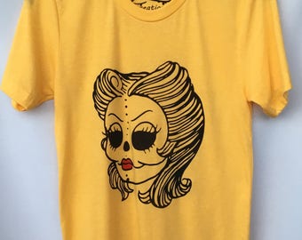 May include: Yellow t-shirt with a black and white illustration of a woman with a skull face and styled hair. The text "W! Creation" is printed on the shirt.