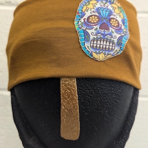 Puede incluir: Una diadema de color amarillo mostaza con un diseño de calavera de azúcar colorido. La calavera tiene ojos azules, una nariz de diamante y un fondo blanco.