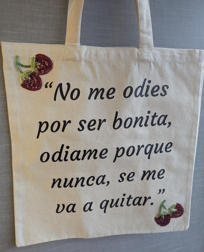 Może przedstawiać: Kremowa torba typu tote z czarnym tekstem: "No me odies por ser bonita, odiame porque nunca, se me va a quitar." Ozdobiona wzorami wiśni z koralik&oacute;w. Posiada dwa uchwyty do przenoszenia.