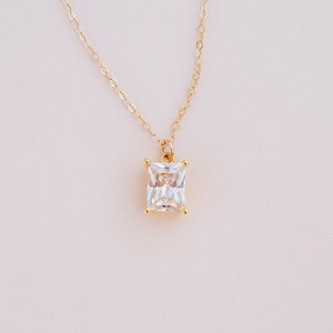 May include: A gold chain necklace with a square-shaped clear crystal pendant.