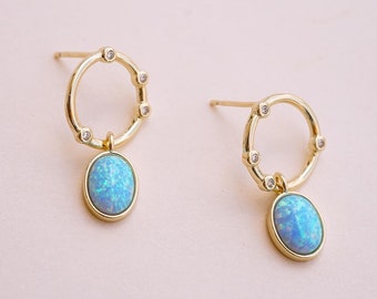 May include: A pair of gold-colored earrings. Each earring features a circular design with small, clear stones and a dangling, oval-shaped, light blue opal. The earrings are set against a soft pink background.
