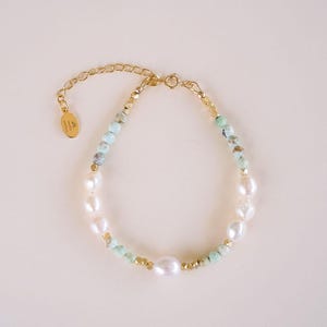 May include: A delicate gold bracelet featuring a mix of white pearls, light blue and yellow beads. The bracelet has a gold chain and clasp, and a small gold charm. The jewelry is displayed on a light background.
