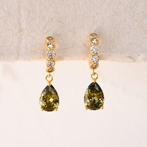 May include: A pair of gold-toned earrings. Each earring features a hoop with small, clear stones and a teardrop-shaped, olive-green gemstone pendant. The earrings are displayed against a neutral background.