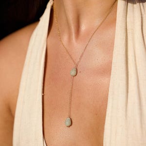 May include: A gold-colored necklace with a delicate chain and two teardrop-shaped, pale green gemstones. The necklace hangs in a Y-shape, with one gemstone at the base of the Y and the other near the neckline. The necklace is a piece of jewelry.
