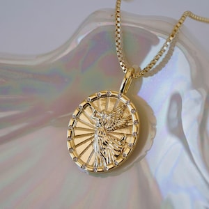 Puede incluir: Collar con colgante dorado con un relieve detallado de una figura alada dentro de un marco circular. El colgante est&aacute; adornado con peque&ntilde;as gemas transparentes alrededor del borde y cuelga de una delicada cadena. El collar se exhibe sobre una superficie nacarada.
