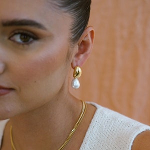 May include: Gold and pearl drop earrings. The earrings feature a gold teardrop-shaped pendant with a pearl hanging from the bottom.