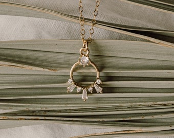 May include: A gold necklace with a delicate circle pendant adorned with small sparkling stones. The necklace is displayed against a backdrop of green palm leaves.