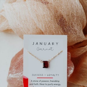 May include: A white card with the text "JANUARY Garnet" and "SUCCESS + LOYALTY" printed on it. A gold chain necklace with a red garnet gemstone pendant is displayed on the card. The text below the gemstone reads "A stone of passion, friendship and faith. Wear to purify energy, bring serenity, and aid in personal and career growth. @WANDERANDLUST JEWELRY"