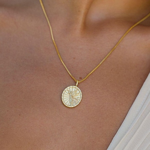 Puede incluir: Un collar dorado con un colgante circular con un dise&ntilde;o detallado. El colgante cuelga de una cadena fina. La persona lleva una blusa blanca plisada.
