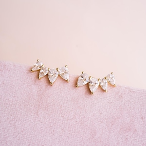 May include: A pair of gold-toned earrings, each featuring three pear-shaped, clear gemstones. The earrings are arranged in a curved line, with the gemstones graduating in size. The background is a soft pink fabric.