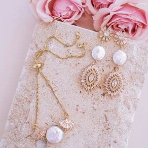 May include: A gold chain bracelet with a pearl charm and a delicate crystal design. The bracelet is displayed on a light beige stone surface with pink roses in the background.