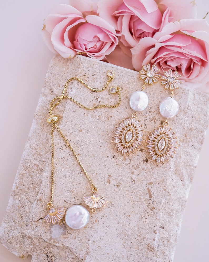 May include: A gold chain bracelet with a pearl charm and a delicate crystal design. The bracelet is displayed on a light beige stone surface with pink roses in the background.