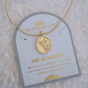 Puede incluir: Collar dorado con un colgante con un dise&ntilde;o de p&aacute;jaro dentro de un marco ovalado. El collar se muestra en una tarjeta que dice "WANDER AND LUST" y "THE GUARDIAN". La tarjeta tambi&eacute;n incluye texto sobre el amor, el consuelo y la paz.